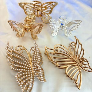 May include: An assortment of butterfly-shaped hair clips in gold and silver tones. The clips feature intricate designs, including openwork, pearl embellishments, and a classic claw clip style. The collection offers a variety of styles for accessorizing.