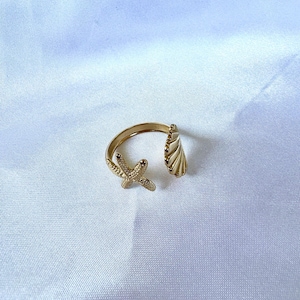 May include: A gold-colored adjustable ring featuring a starfish and a seashell design. The ring is open, allowing for size adjustment. The starfish has textured details, and the seashell has a ribbed pattern. The ring is set against a white background.
