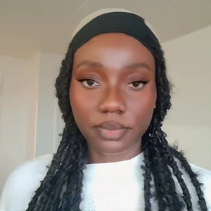 May include: A woman with dark brown skin is wearing a black headband and long black braids. She is wearing a white sweater and looking directly at the camera.