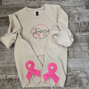 Puede incluir: Una sudadera beige con el texto "Stronger than the Storm" en escritura negra y un lazo rosa. Dos grandes apliques de lazo rosa están en las mangas. La sudadera está colocada sobre una superficie de madera.
