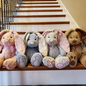 May include: Four plush bunny rabbits with long floppy ears, sitting on a wooden staircase. The bunnies are in different colours: pink, blue, grey, and brown. Each bunny has a name embroidered on one of its ears: "Samantha", "Sawyer", "Riley", and "Junior".