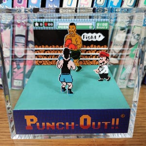 Op de afbeelding: Een helder acrylblok met een diorama van het videospel "Punch-Out!!". De sc&egrave;ne toont gepixelde figuren van boksers en een scheidsrechter, met de spelinterface zichtbaar. De basis van het blok toont de titel van het spel in oranje en blauw.