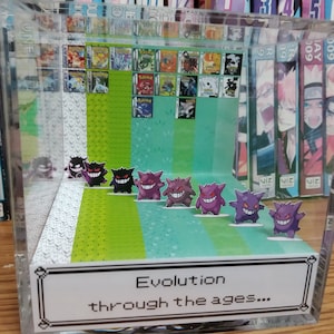 May include: A clear acrylic box with a diorama of the Pokemon Ghost-type evolution line. The figures are arranged in a line from left to right, starting with Gastly, then Haunter, then Gengar. The box has a green and white patterned background and a label that reads "Evolution through the ages..."