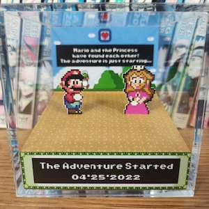 May include: A clear acrylic box containing a miniature diorama. The diorama features pixelated characters from the Super Mario Bros. video game, including Mario and Princess Peach. The diorama is set against a green background with a blue sky. The box has a label on the bottom that reads "The Adventure Started 04'25'2022".