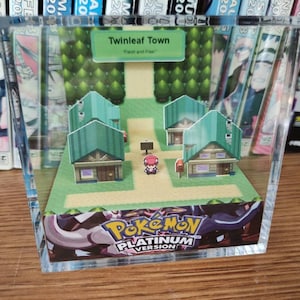 May include: A clear acrylic display featuring a diorama of Twinleaf Town from Pokemon Platinum Version. The scene includes paper cutouts of houses, trees, a character, and the game's logo. The text "Twinleaf Town" and "Fresh and Free" are visible.