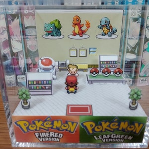 May include: A clear acrylic display case with a miniature diorama of a Pokemon game store. The diorama features a pixelated design with a red-roofed building, a counter with a cash register, and a shelf with Pokemon items. The diorama also includes three pixelated Pokemon characters: Bulbasaur, Charmander, and Squirtle. The case has a red and green base with the text "Pokemon FireRed Version" and "Pokemon LeafGreen Version".