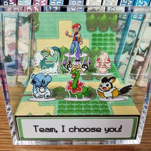 Puede incluir: Una pantalla de acr&iacute;lico transparente con una escena pixelada de personajes de dibujos animados. La escena incluye un entrenador y varias criaturas coloridas, con el texto "Team, I choose you!" en la base. La pantalla est&aacute; colocada frente a una colecci&oacute;n de mangas.