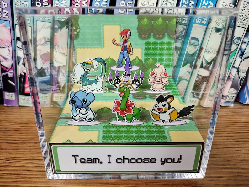 Puede incluir: Una pantalla de acr&iacute;lico transparente con una escena pixelada de personajes de dibujos animados. La escena incluye un entrenador y varias criaturas coloridas, con el texto "Team, I choose you!" en la base. La pantalla est&aacute; colocada frente a una colecci&oacute;n de mangas.
