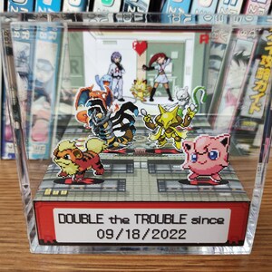 Puede incluir: Una vitrina de acr&iacute;lico transparente con un diorama 3D de personajes Pok&eacute;mon. El diorama presenta una variedad de Pok&eacute;mon, incluyendo Charmeleon, Arcanine, Jolteon y Jigglypuff. La vitrina tiene una etiqueta roja y blanca que dice "DOUBLE the TROUBLE since 09/18/2022".