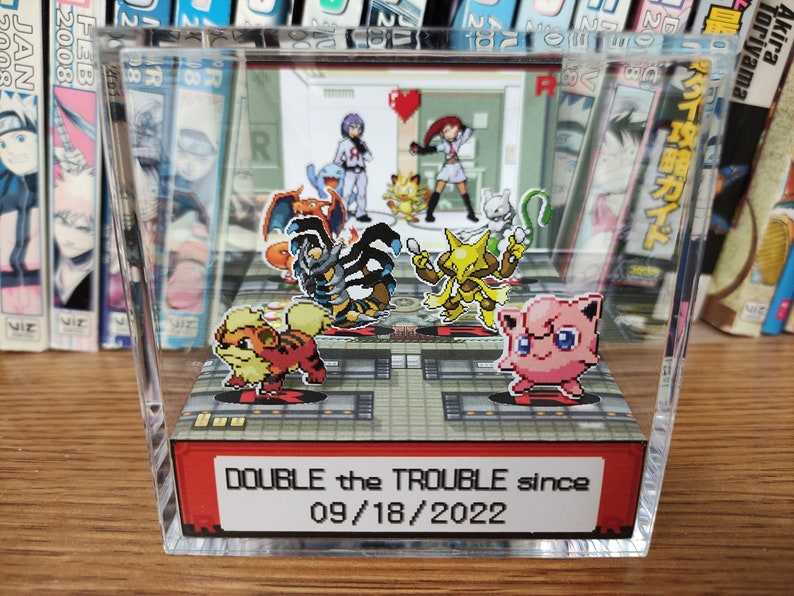 Puede incluir: Una vitrina de acr&iacute;lico transparente con un diorama 3D de personajes Pok&eacute;mon. El diorama presenta una variedad de Pok&eacute;mon, incluyendo Charmeleon, Arcanine, Jolteon y Jigglypuff. La vitrina tiene una etiqueta roja y blanca que dice "DOUBLE the TROUBLE since 09/18/2022".