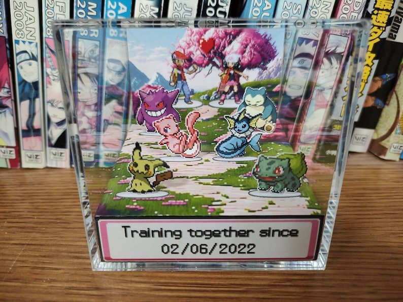 May include: A clear acrylic display case with a 3D diorama of Pokemon characters. The diorama features a pink and white background with a cherry blossom tree. The characters include Pikachu, Bulbasaur, Charmander, Squirtle, and a few others. The case has a pink label with the text "Training together since 02/06/2022".