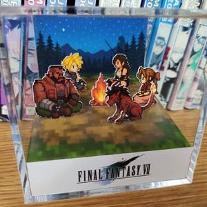 May include: A clear acrylic display featuring a pixelated scene from Final Fantasy VII. The scene depicts characters around a campfire under a starry night sky. The Final Fantasy VII logo is at the bottom.