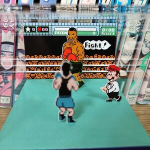 Op de afbeelding: Een diorama met een retro videogame sc&egrave;ne met gepixelde karakters in een boksring. De karakters zijn onder meer een bokser, een Mario-achtig figuur en een andere bokser. De achtergrond bevat mangaboeken.