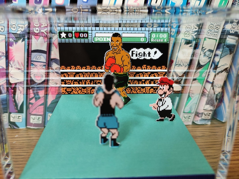 Op de afbeelding: Een diorama met een retro videogame sc&egrave;ne met gepixelde karakters in een boksring. De karakters zijn onder meer een bokser, een Mario-achtig figuur en een andere bokser. De achtergrond bevat mangaboeken.