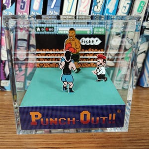 Op de afbeelding: Een helder acrylblok met een miniatuur diorama van het videospel "Punch-Out!!". De sc&egrave;ne toont gepixelde personages in een boksring, met de titel van het spel. Op de achtergrond is een plank met manga's te zien.