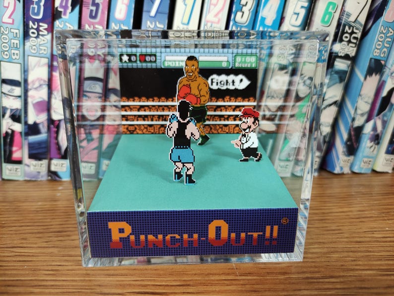 Op de afbeelding: Een helder acrylblok met een miniatuur diorama van het videospel "Punch-Out!!". De sc&egrave;ne toont gepixelde personages in een boksring, met de titel van het spel. Op de achtergrond is een plank met manga's te zien.