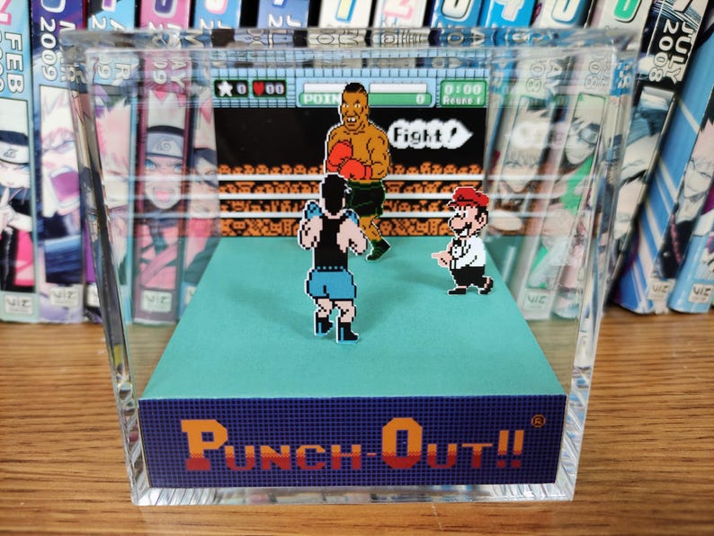 Op de afbeelding: Een helder acrylblok met een diorama van het videospel "Punch-Out!!". De sc&egrave;ne toont gepixelde figuren van boksers en een scheidsrechter, met de spelinterface zichtbaar. De basis van het blok toont de titel van het spel in oranje en blauw.