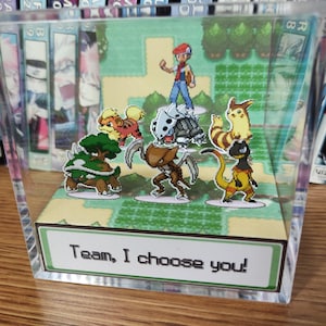 Puede incluir: Una pantalla acr&iacute;lica transparente con un diorama de una escena de videojuego con personajes de dibujos animados. La escena incluye un entrenador y varias criaturas coloridas. El texto "Team, I choose you!" se muestra en la parte inferior.