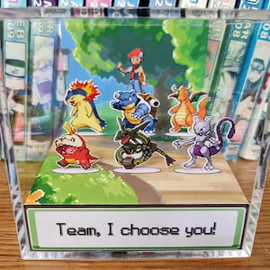 Puede incluir: Un bloque acr&iacute;lico transparente con un diorama colorido de personajes de Pok&eacute;mon. La escena incluye un entrenador y varios Pok&eacute;mon en un camino, con el texto "Team, I choose you!" en la base. El fondo muestra un paisaje verde.