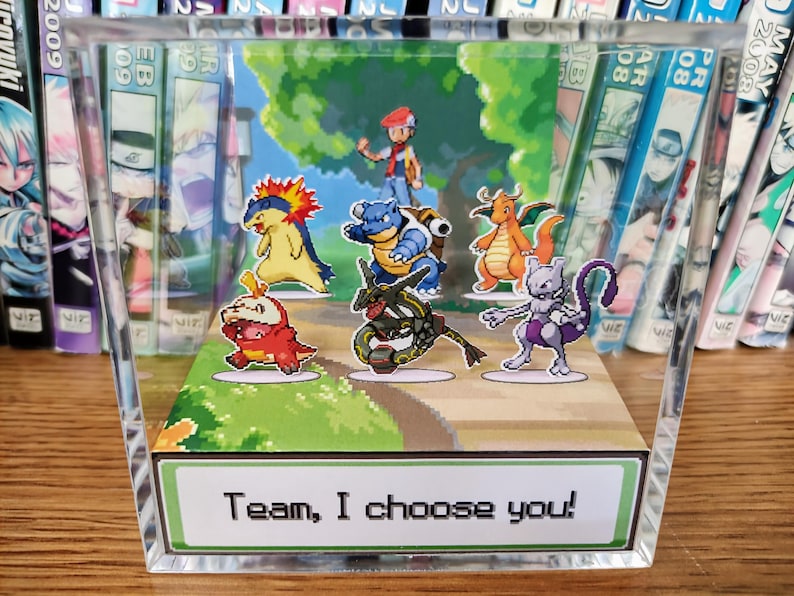 Puede incluir: Un bloque acr&iacute;lico transparente con un diorama colorido de personajes de Pok&eacute;mon. La escena incluye un entrenador y varios Pok&eacute;mon en un camino, con el texto "Team, I choose you!" en la base. El fondo muestra un paisaje verde.