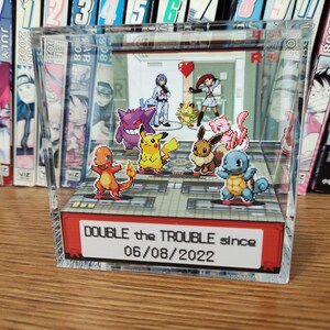 Puede incluir: Una vitrina de cubo de acr&iacute;lico transparente con un diorama de personajes de Pok&eacute;mon en su interior. Los personajes est&aacute;n dispuestos en un suelo cuadriculado con un cartel rojo que dice "DOUBLE the TROUBLE since 06/08/2022".