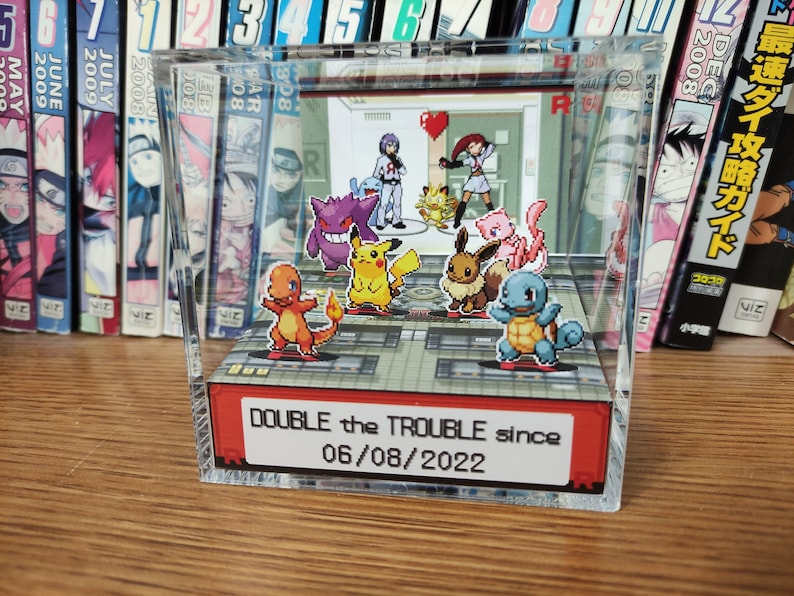 Puede incluir: Una vitrina de cubo de acr&iacute;lico transparente con un diorama de personajes de Pok&eacute;mon en su interior. Los personajes est&aacute;n dispuestos en un suelo cuadriculado con un cartel rojo que dice "DOUBLE the TROUBLE since 06/08/2022".