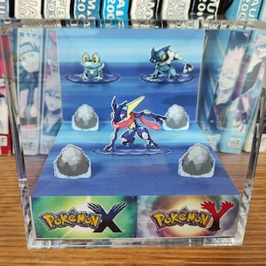 May include: A clear acrylic cube display case with a 3D papercraft scene of Pokemon characters from the Pokemon X and Y video games. The scene features a blue background with water, rocks, and three Pokemon: Greninja, Froakie, and Delphox.