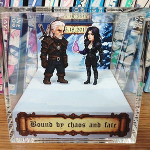 May include: A pixelated diorama featuring two characters from a fantasy game, standing on a white platform inside a clear acrylic box. The text "Bound by chaos and fate" is displayed on a banner. The background shows a snowy landscape.