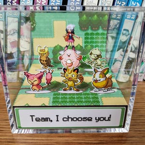 Puede incluir: Una pantalla acr&iacute;lica transparente con un diorama de una escena de videojuego con varios personajes de dibujos animados. La escena incluye un entrenador y varias criaturas coloridas. El texto "Team, I choose you!" se muestra en una etiqueta verde y blanca.