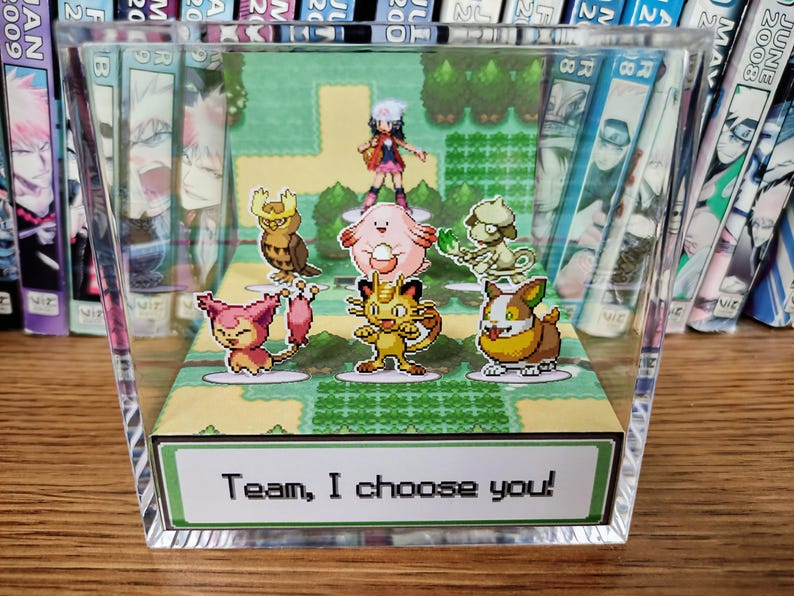 Puede incluir: Una pantalla acr&iacute;lica transparente con un diorama de una escena de videojuego con varios personajes de dibujos animados. La escena incluye un entrenador y varias criaturas coloridas. El texto "Team, I choose you!" se muestra en una etiqueta verde y blanca.