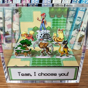 Puede incluir: Bloque acr&iacute;lico transparente con un diorama de videojuego colorido. Presenta personajes de dibujos animados y un paisaje verde. El texto "Team, I choose you!" est&aacute; en la parte inferior.