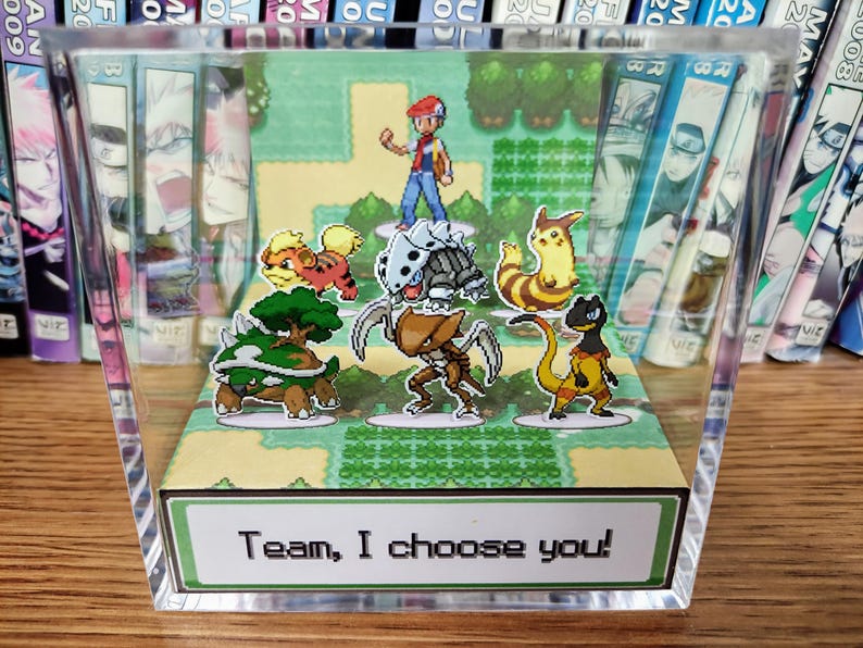 Puede incluir: Bloque acr&iacute;lico transparente con un diorama de videojuego colorido. Presenta personajes de dibujos animados y un paisaje verde. El texto "Team, I choose you!" est&aacute; en la parte inferior.