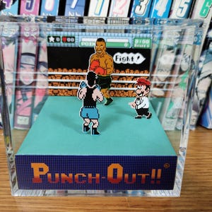 Op de afbeelding: Een heldere acryl display case met een gepixeleerde bokssc&egrave;ne uit het videospel "Punch-Out!!". De sc&egrave;ne bevat twee boksers in een ring, met Mario als scheidsrechter. De basis van de case toont de titel van het spel in oranje en blauw.