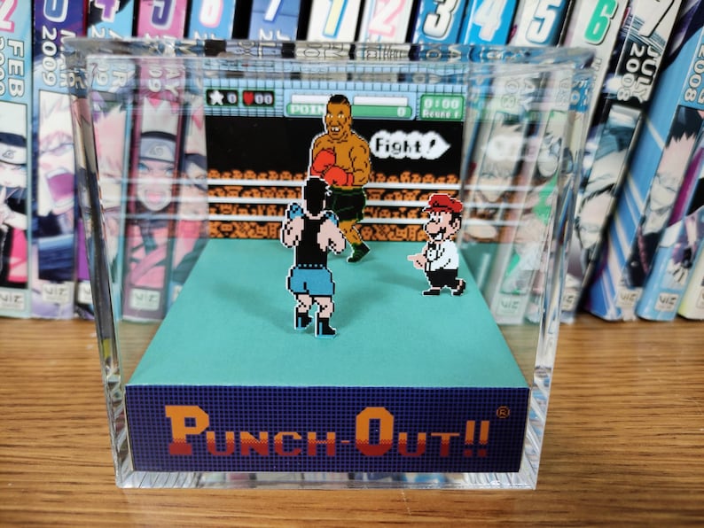 Op de afbeelding: Een heldere acryl display case met een gepixeleerde bokssc&egrave;ne uit het videospel "Punch-Out!!". De sc&egrave;ne bevat twee boksers in een ring, met Mario als scheidsrechter. De basis van de case toont de titel van het spel in oranje en blauw.