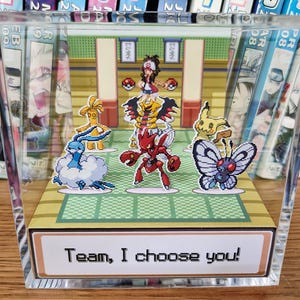 Puede incluir: Un bloque acr&iacute;lico transparente con un diorama colorido de una escena de batalla Pok&eacute;mon. La escena incluye varios personajes Pok&eacute;mon y un entrenador, con el texto "Team, I choose you!" en la parte inferior. El bloque se muestra frente a una estanter&iacute;a.