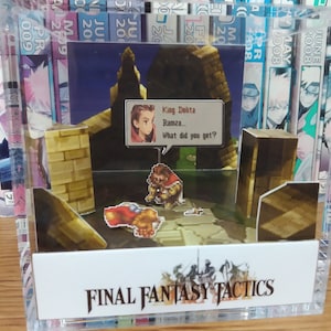 May include: A clear plastic display case with a 3D diorama of a scene from the video game Final Fantasy Tactics. The diorama features characters from the game, including Ramza and King Delita, standing on a stone path. The text "FINAL FANTASY TACTICS" is printed on the base of the display case.