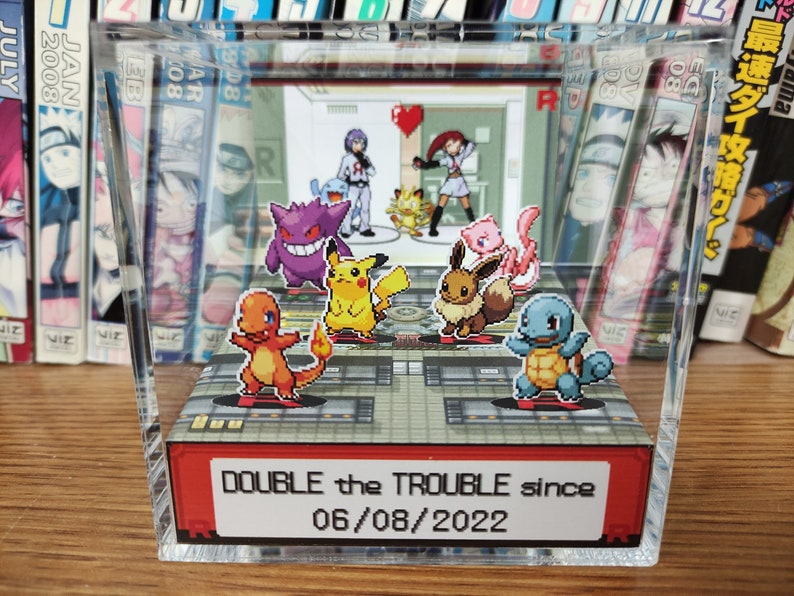 Puede incluir: Una vitrina de acr&iacute;lico transparente con un diorama 3D de personajes de Pok&eacute;mon. El diorama presenta a Pikachu, Squirtle, Bulbasaur y otros. La vitrina tiene una base roja con el texto "DOUBLE the TROUBLE since 06/08/2022".