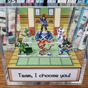Puede incluir: Un bloque acr&iacute;lico transparente con un colorido diorama de una escena de batalla Pok&eacute;mon. La escena incluye varios personajes Pok&eacute;mon y un entrenador. El texto "Team, I choose you!" se muestra en la parte inferior. El bloque se exhibe sobre una superficie de madera.