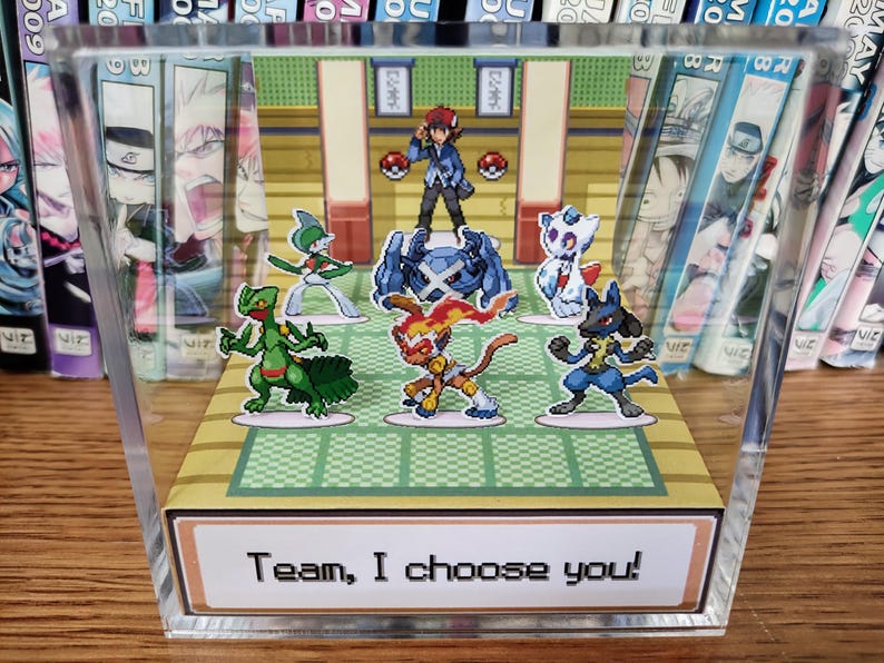 Puede incluir: Un bloque acr&iacute;lico transparente con un colorido diorama de una escena de batalla Pok&eacute;mon. La escena incluye varios personajes Pok&eacute;mon y un entrenador. El texto "Team, I choose you!" se muestra en la parte inferior. El bloque se exhibe sobre una superficie de madera.