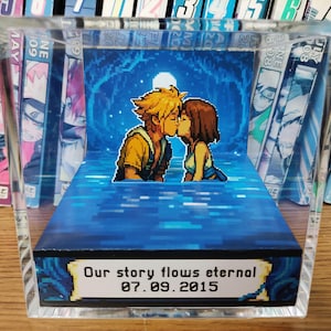 May include: A pixelated diorama featuring a couple kissing under a full moon, set against a blue background. The base reads "Our story flows eternal 07.09.2015". The scene is encased in a clear acrylic block, with manga volumes visible on either side.