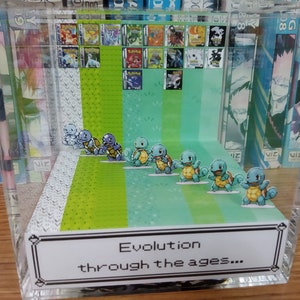 May include: A clear plastic display case with a diorama of the evolution of Squirtle from Pokemon. The diorama includes a series of Squirtle figurines, each representing a different stage of evolution. The case also includes a background with a green and blue pattern, and a label that reads "Evolution through the ages..."