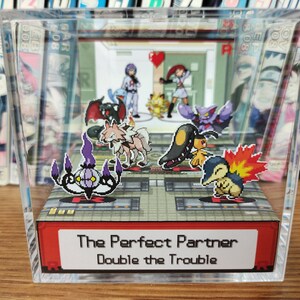 Puede incluir: Una vitrina de cubo de acr&iacute;lico transparente con una base roja. La vitrina presenta un diorama de personajes de Pok&eacute;mon, incluyendo un Gengar morado y negro, un Gardevoir blanco y marr&oacute;n, un Flareon marr&oacute;n y amarillo, un Misdreavus morado y negro, un Espeon marr&oacute;n y amarillo y un Ampharos verde y amarillo. La base de la vitrina tiene el texto "The Perfect Partner Double the Trouble".