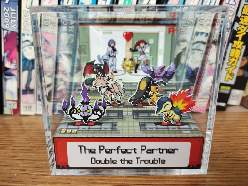 Puede incluir: Una vitrina de cubo de acr&iacute;lico transparente con una base roja. La vitrina presenta un diorama de personajes de Pok&eacute;mon, incluyendo un Gengar morado y negro, un Gardevoir blanco y marr&oacute;n, un Flareon marr&oacute;n y amarillo, un Misdreavus morado y negro, un Espeon marr&oacute;n y amarillo y un Ampharos verde y amarillo. La base de la vitrina tiene el texto "The Perfect Partner Double the Trouble".