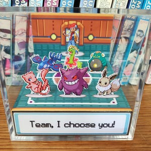 Puede incluir: Una vitrina de acr&iacute;lico transparente con una escena pixelada de Pok&eacute;mon. Presenta un entrenador y varios personajes Pok&eacute;mon. El texto "Team, I choose you!" est&aacute; en la base. El fondo muestra libros manga.