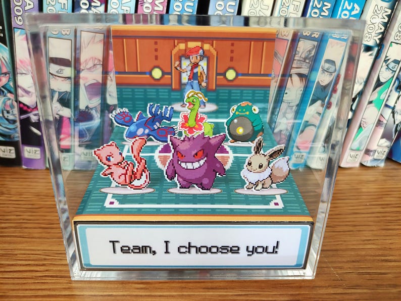 Puede incluir: Una vitrina de acr&iacute;lico transparente con una escena pixelada de Pok&eacute;mon. Presenta un entrenador y varios personajes Pok&eacute;mon. El texto "Team, I choose you!" est&aacute; en la base. El fondo muestra libros manga.