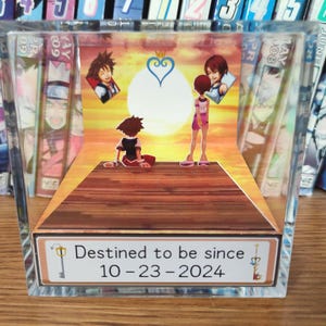 Puede incluir: Un bloque acrílico transparente con un diorama 3D que representa personajes de anime en un muelle de madera contra un atardecer. El diorama incluye el texto "Destined to be since 10-23-2024". Se ven libros de manga en el fondo.