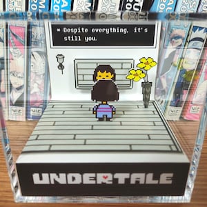 May include: A clear acrylic display featuring a pixelated scene from the video game Undertale. The scene includes a character, text that reads "Despite everything, it's still you," and a stylized background. The base of the display reads "UNDERTALE."