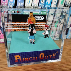 Op de afbeelding: Een heldere acryl display met een diorama van het videospel "Punch-Out!!". De sc&egrave;ne toont gepixelde personages in een boksring, met de titel van het spel in oranje en blauw. Op de achtergrond is een plank met kleurrijke manga te zien.