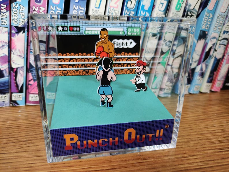 Op de afbeelding: Een heldere acryl display met een diorama van het videospel "Punch-Out!!". De sc&egrave;ne toont gepixelde personages in een boksring, met de titel van het spel in oranje en blauw. Op de achtergrond is een plank met kleurrijke manga te zien.