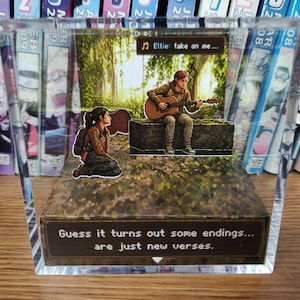 May include: A clear acrylic block featuring a scene with two figures, one playing a guitar. The text "Ellie: take on me..." and "Guess it turns out some endings... are just new verses" are visible. The background includes a forest scene and a bookshelf.